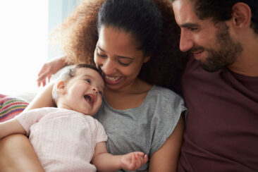 Parents Sitting On Sofa Cuddling Baby Daughter At Home