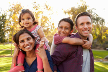 Mixed race parents carry their kids piggyback in a park